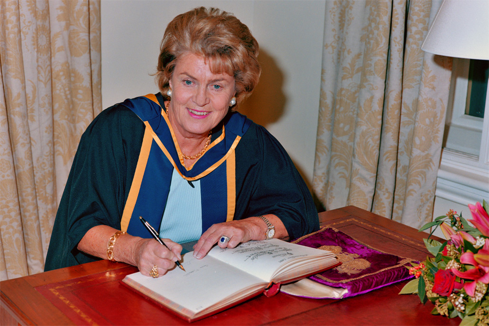 A lady with red hair wearing an academic gown and signing a book on a desk